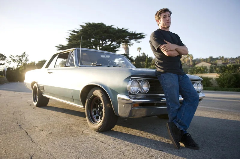 A young man leaning against his vintage muscle car at sunset