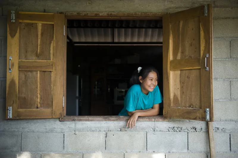 A girl smiling from an open wooden window in a village, northern Thailand